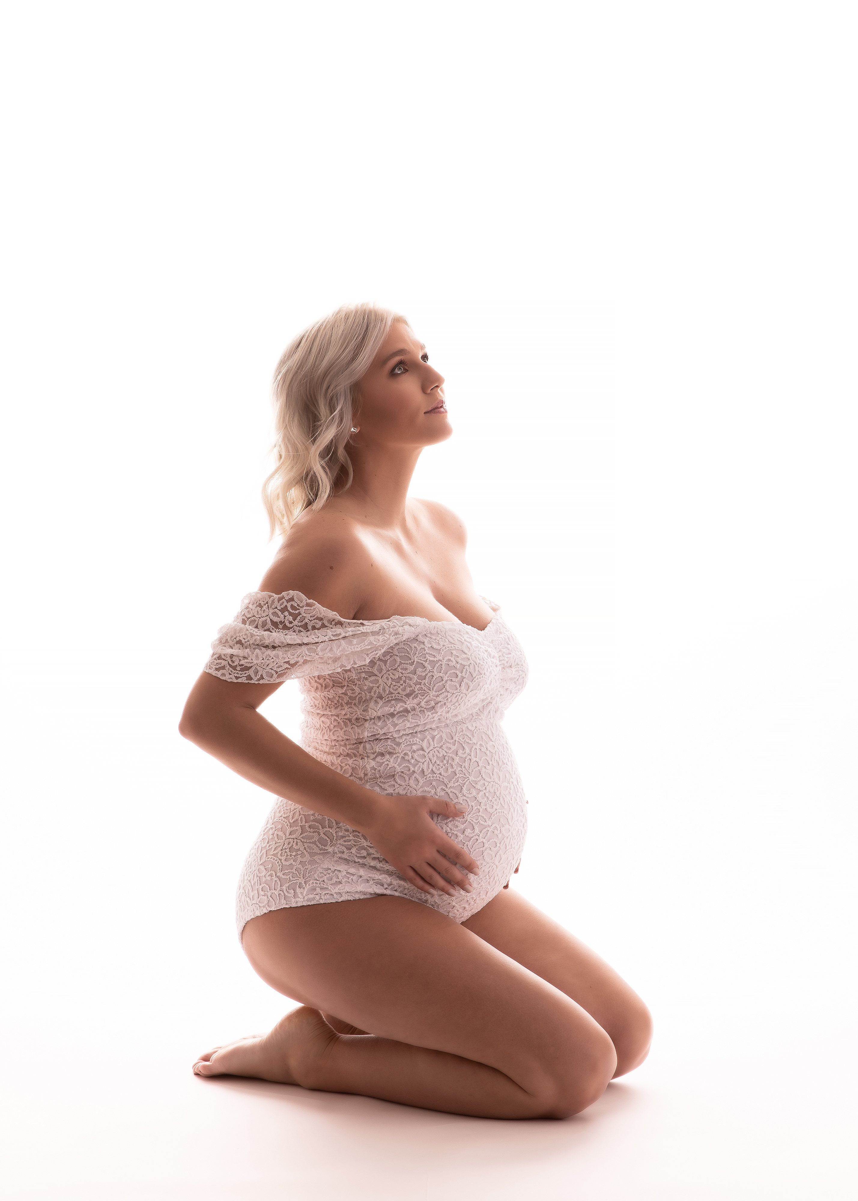 Maternity Photographer, an expectant mother wears a lacy body suit and holds her belly as she sits on her knees