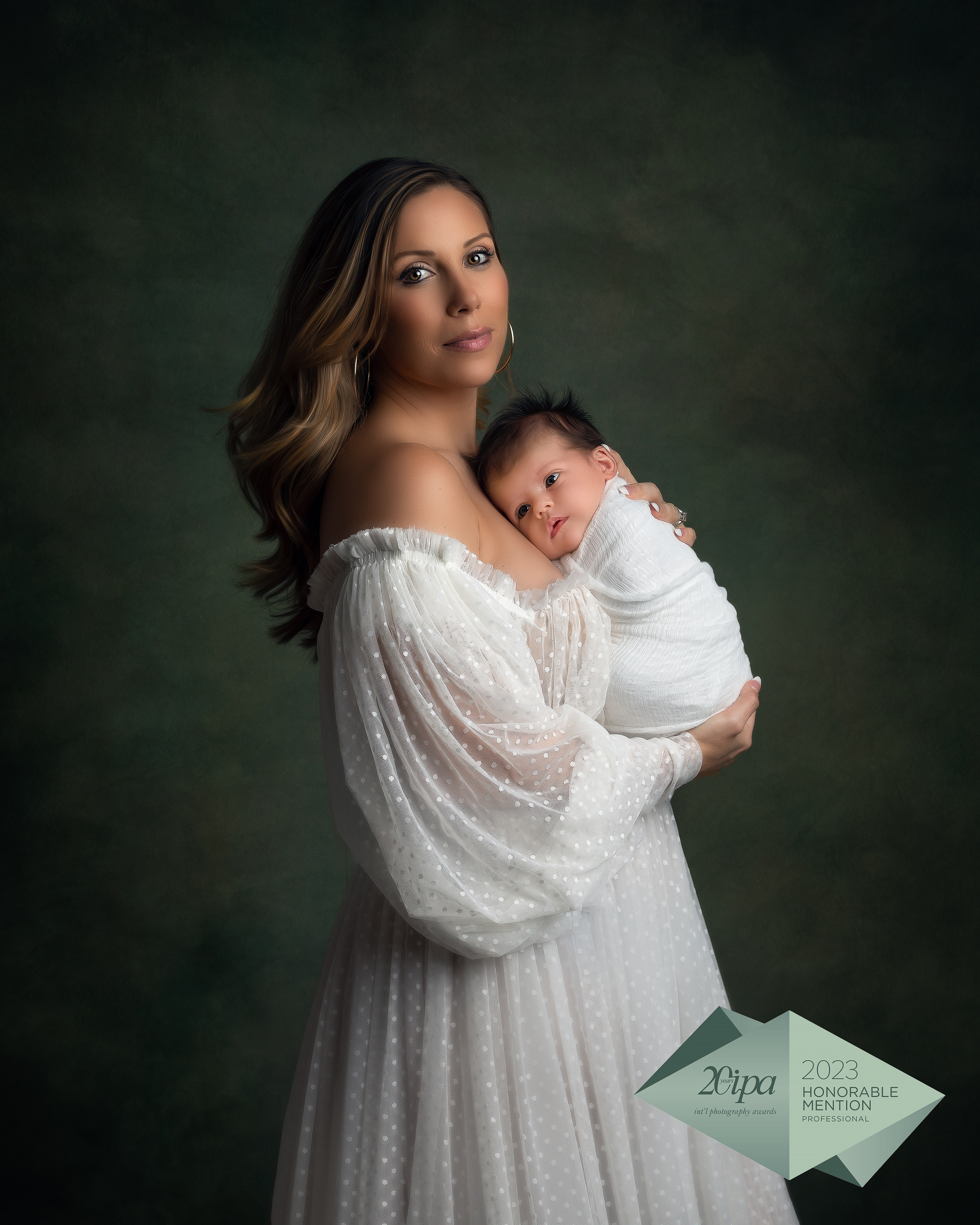 Newborn Photographer, a mother wears a white dress and holds her baby against a dark green studio backdrop