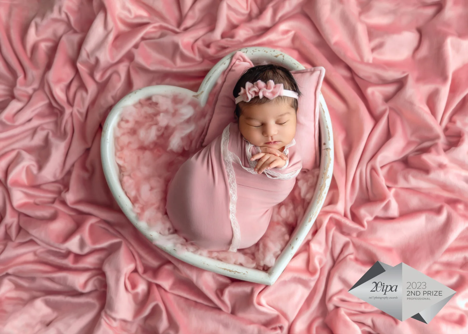 Newborn Photographer, a baby girl sits in a small wooded basket shaped as a heart, she is surrounded by pink linens, a badge reads "2Cipa 2023 2nd prize"